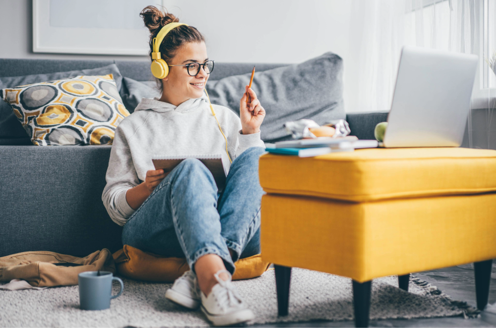 Young woman wearing yellow headphones sits on floor of living room across from laptop