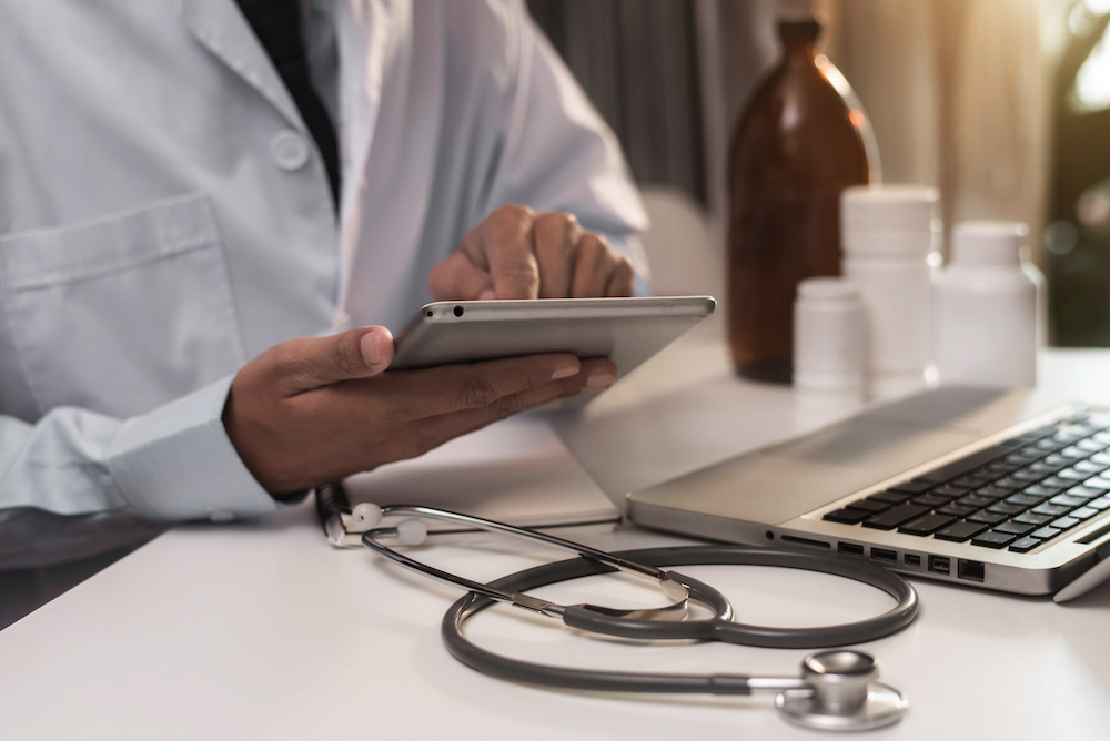 Doctor using tablet at desk with stethoscope, medication bottles, and laptop